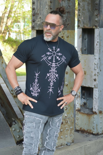 The Ancient Tribal Dude wearing a black t-shirt with white designs standing on a metal bridge.