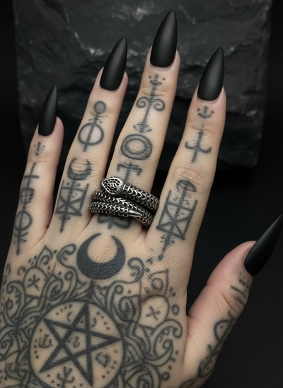 Hand with tattoos and black nail polish wearing an Ancient Tribal triple wrap snake ring against a dark background